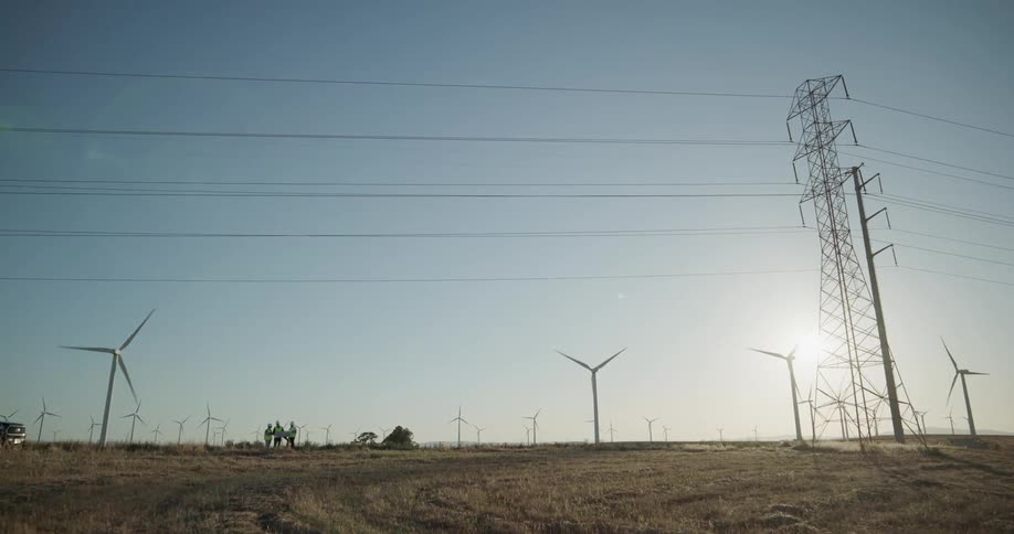Workers stand near on wind farm discussing plans as sun shines past wind turbines