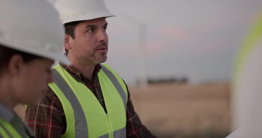 Three construction workers discuss plans as they stand in a field of wind turbines