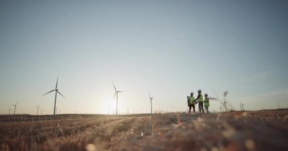 Engineers wearing white hard hats and green vests check blueprints near the wind turbines at sunset