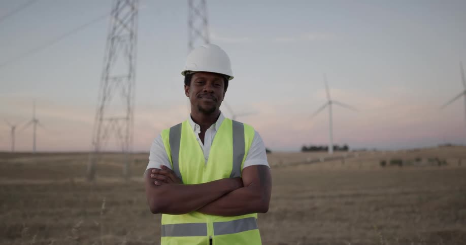 Construction worker wearing hard hat and yellow vest stands in front of transmission towers and wind turbines at sunset