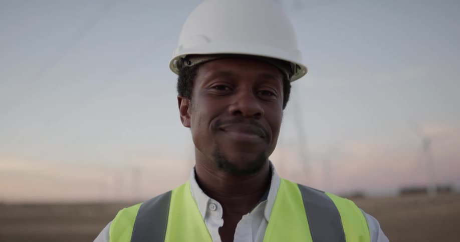 A man in a hard hat and safety vest smiles into the camera while standing on a wind farm in the evening