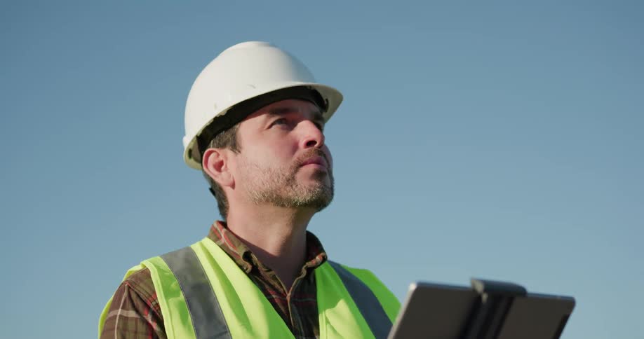 A caucasian male ina hard hat holding a clip board outside looking around