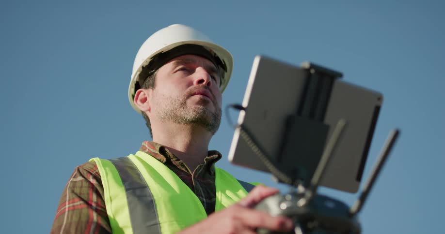 Supervisor wearing a white hardhat and green vest remote controls a drone over a construction site