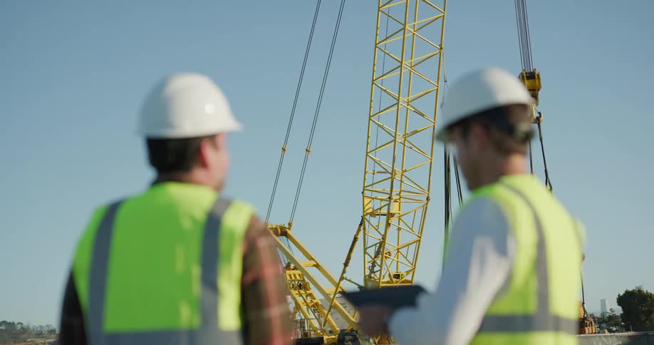 Supervisors wearing white hard hats and green vests fly a drone over a construction site