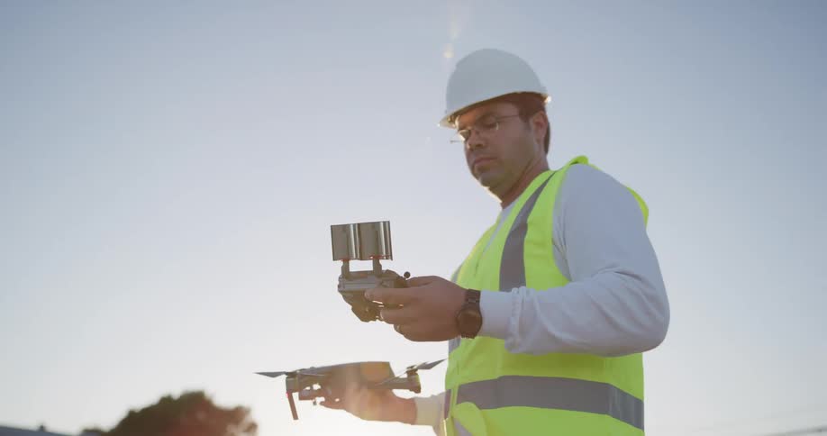 A man in a safety vest and hard hat stands outdoors while holding a drone in one hand and its remote controller in the other