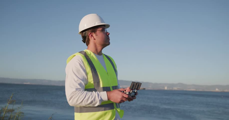 A man in a hard hat and vest stands in front of a body of water and uses a scientific device