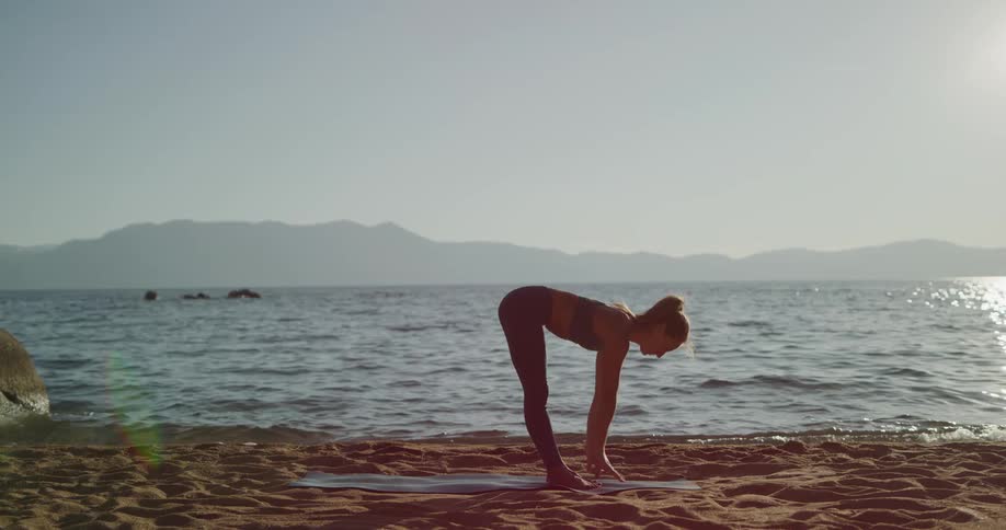 Fit woman practices yoga on a mat over the sand on the coast of a lake in the mountains at sunset
