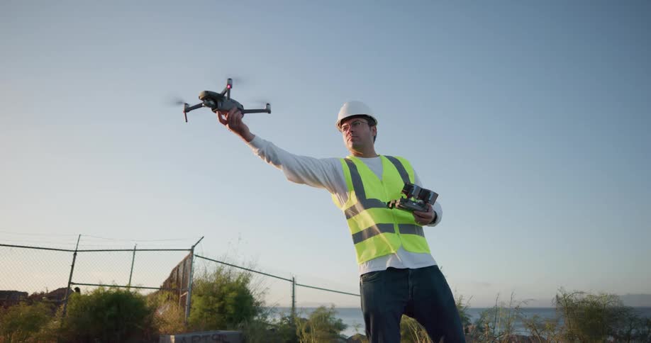 A man wearing a construction vest and hard hat holds a controller for a drone flying in front of him