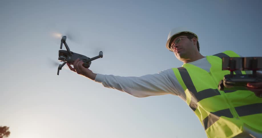 Man wearing a hard hat and safety vest holds a controller as he reaches for a flying drone