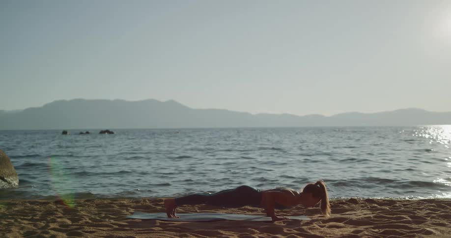 Fit woman practicing yoga in a mat over the sand in the shore of a lake on the mountains at sunset