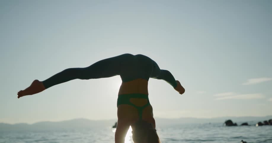 A woman doing a handstand on the shore and stretching with the sky and clouds in the background