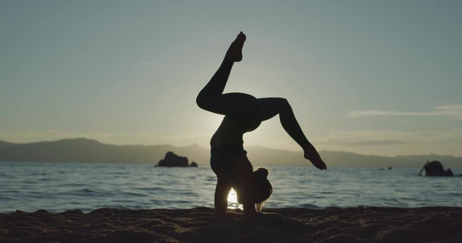 A woman doing a handstand and stretching on the shore with the sky and clouds in the background