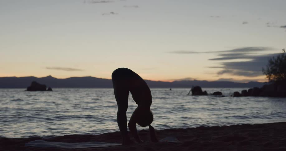 Young woman practices yoga at the shore of a lake in the mountains at sunset