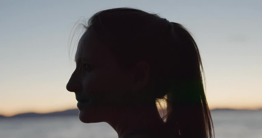 Young blonde woman stands meditatively on the lake shore near the mountains at sunset