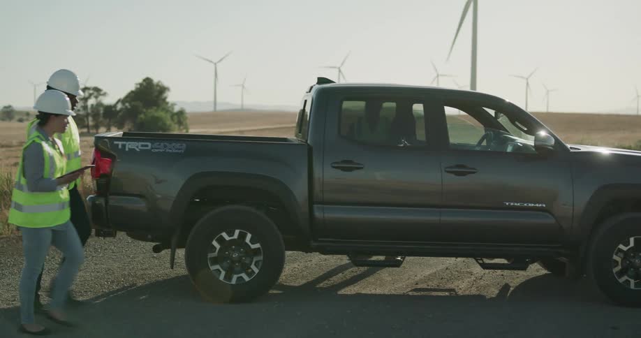 A group of two men and a woman enter a truck parked on a gravel road that runs through a field of wind turbines during the day