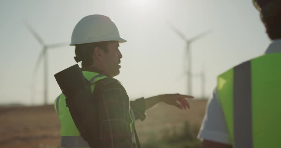 Two men and a woman in safety vests and hard hats walk down a dirt road through a field of wind turbines
