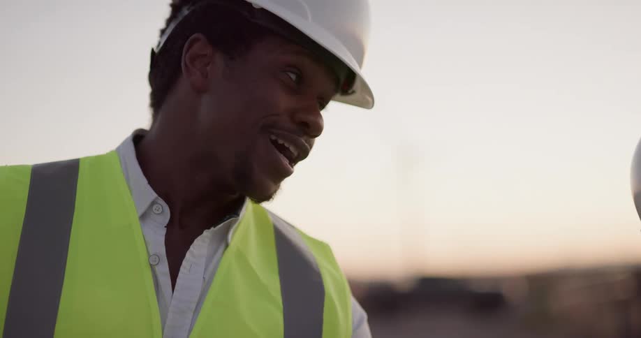 Engineers wearing white hard hats and green vests study a blueprint near wind turbines at sunset
