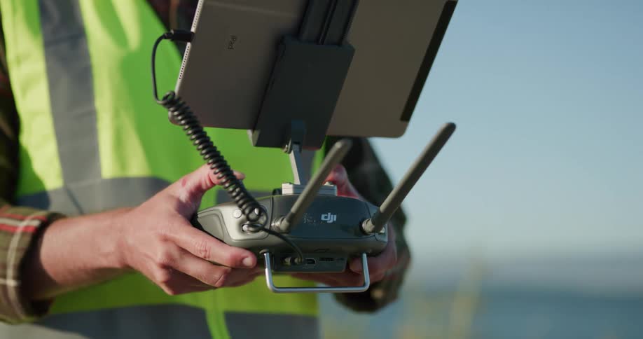 A man in a safety vest stands outdoors during the day holding a radio drone controller with a tablet plugged into it