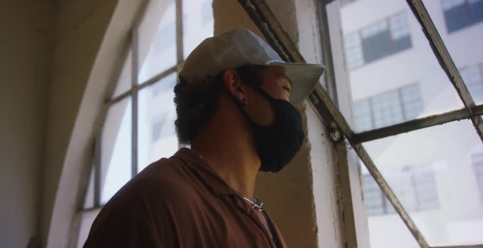 Asian man with a cap and a black mask looks out the window