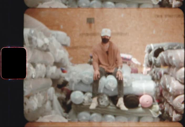 Man wearing face mask and baseball cap sits on rolled bundles stacked and stored in building