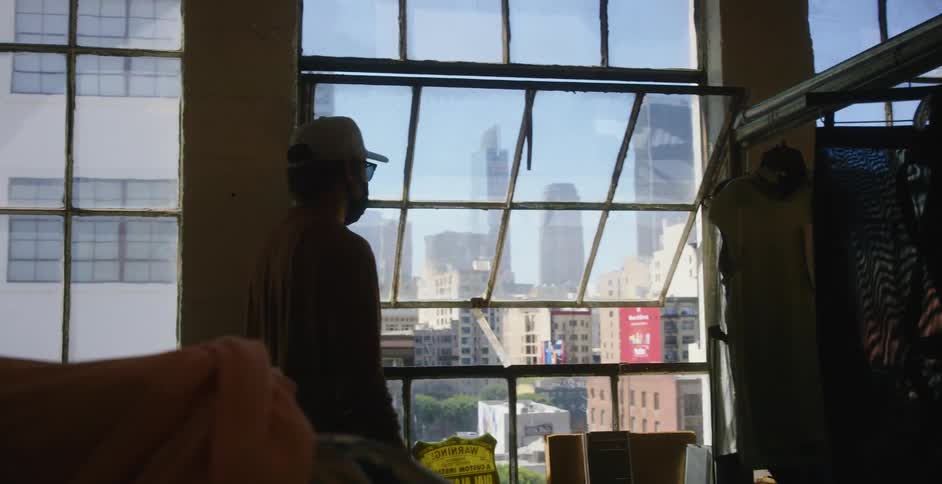 Man walks past clothes hanging in open to look out open window at downtown skyscrapers