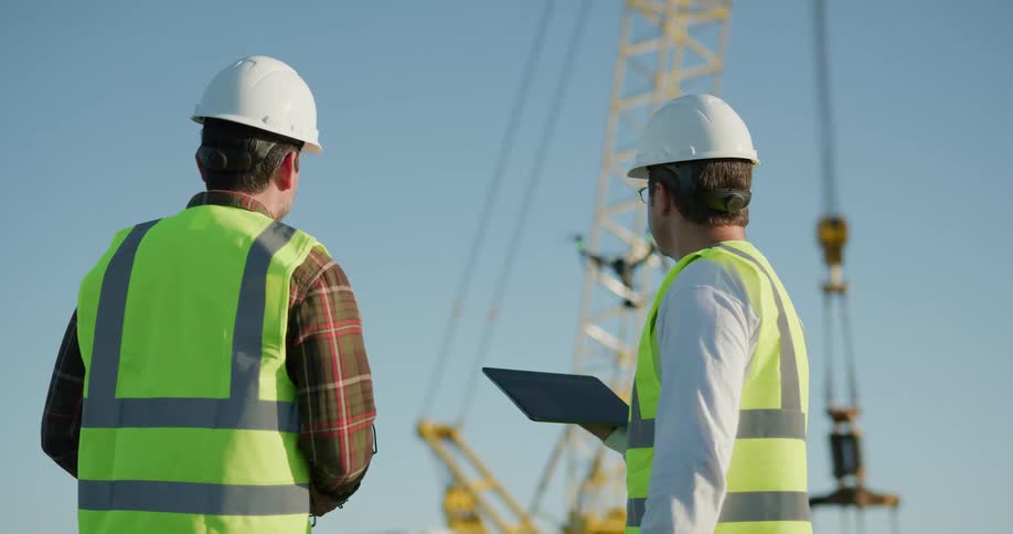 Two men wearing vests and hard hats standing outside operating a drone