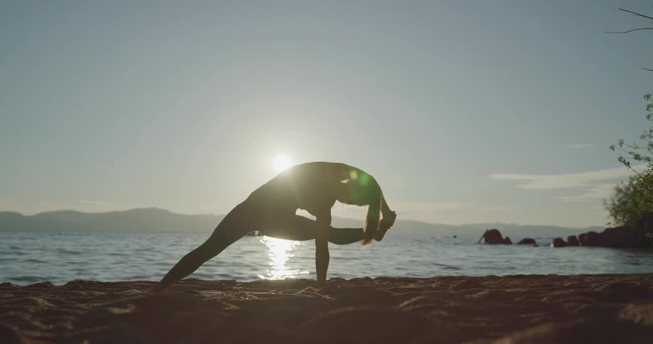 A woman bends over in a yoga pose standing on the beach with a low sun in the background