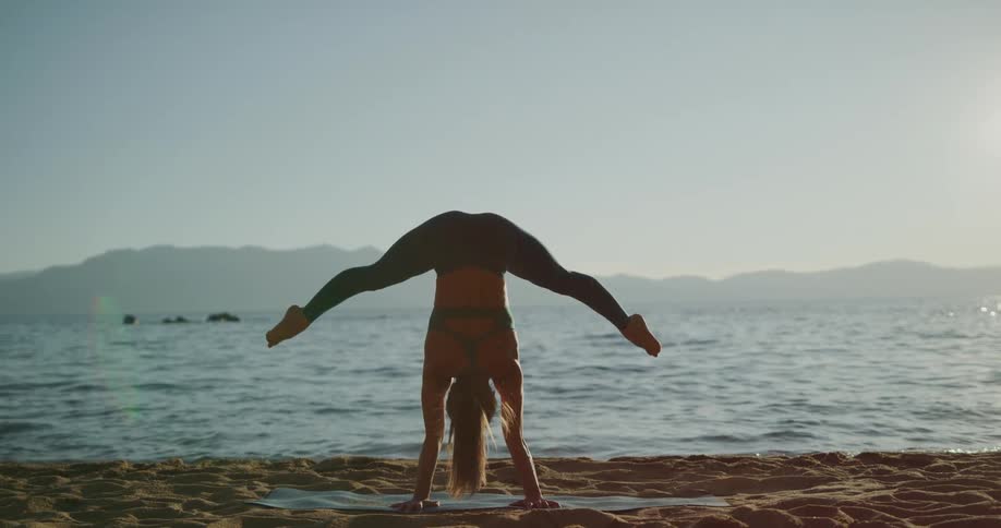 A woman on the beach doing a handstand and stretching with the water and the sky in the background
