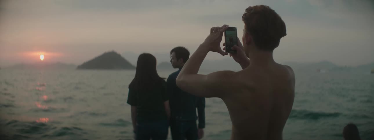 A man and woman gaze at one another while standing together on the coast at sunset as a shirtless man takes photos of them from behind