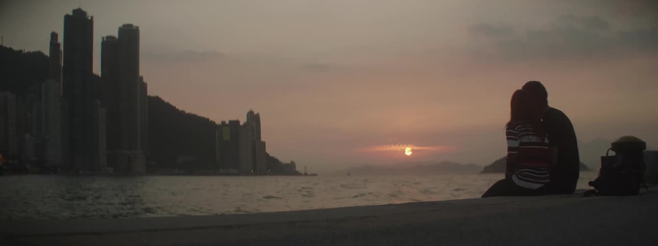 A man and woman sit closely together on the coast at sunsest as they look out over the bay towards the skyscrapers of the city on the other side