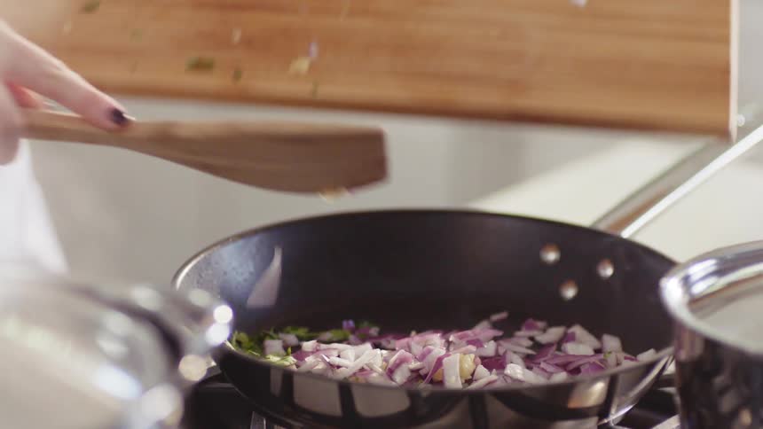 A cook uses a wooden spatula to push chopped vegetables from a cutting board into a skillet