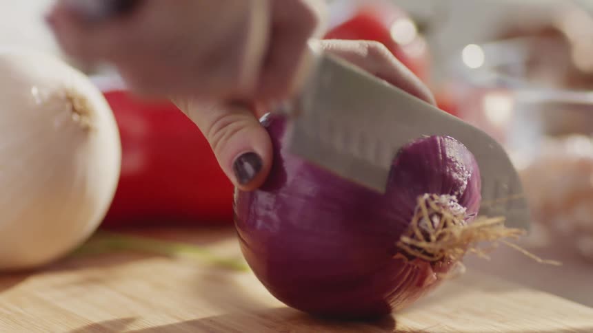 A woman uses a knife to slice the end off of a red onion on a wooden cutting board