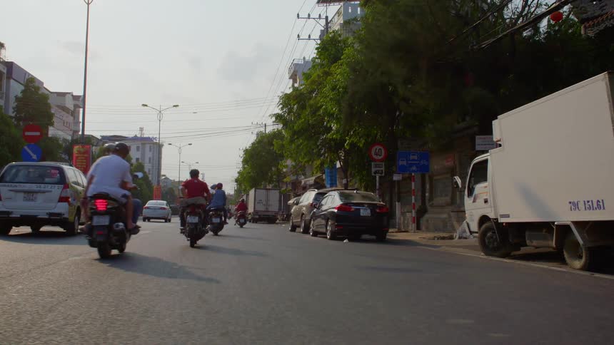Motorists travel along street past buildings and parked vehicles