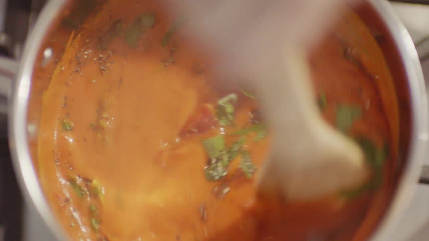 A person stirring a pot of sauce cooking on a stove top with a wood spoon