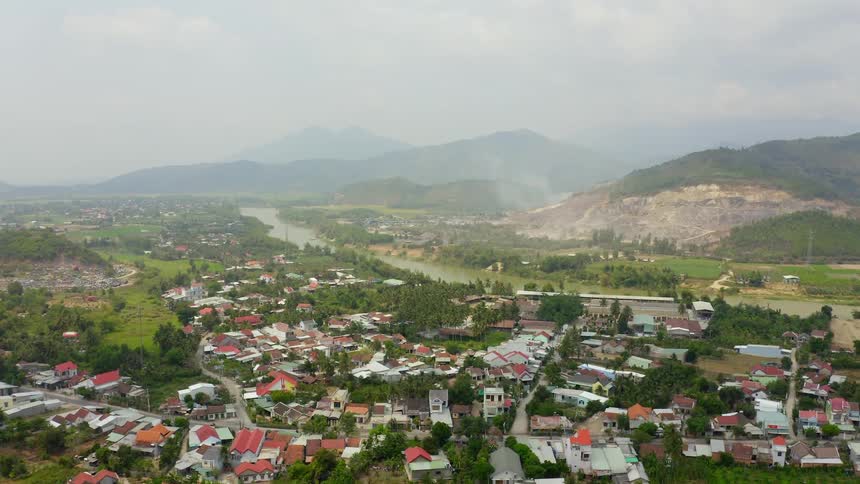 A small town bordering a murky river below a group of mountains shrouded in fog