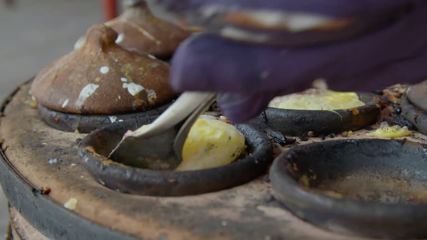 A person wearing a glove using a small spatula to unstick a bit of food from the small pan it is cooking in