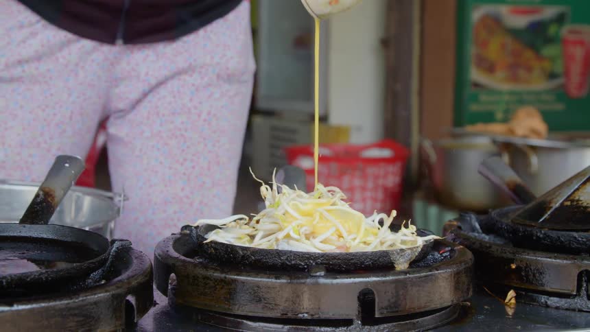 A person pours whisked eggs over a pile of bean sprouts on a grill
