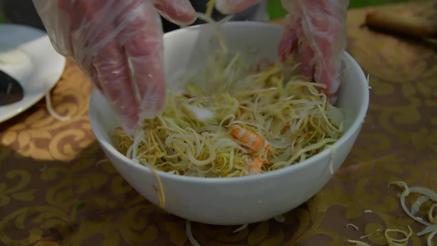 A person with gloves mixing a bowl of noodles and shrimp on a table