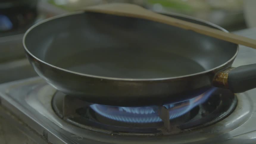 A frying pan and wooden spatula sit on a stovetop as the gas burner comes on