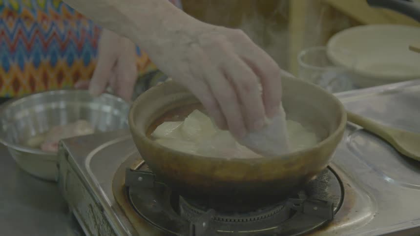 An elderly woman cooks some chicken in a bowl over a burner