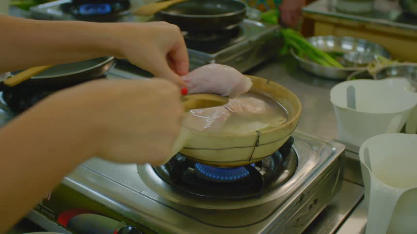 A chef places a piece of raw chicken into a pot of water on a stove in a professional kitchen