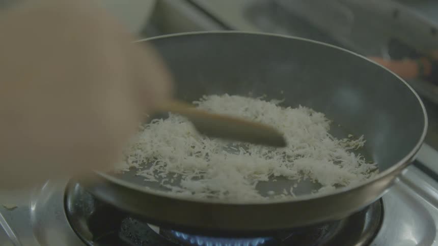 A person stirs shredded cheese in a frying pan on an oven burner