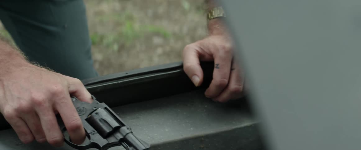 A man with glasses and a wristwatch holding a gun while looking into a car trunk