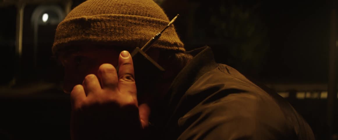 Man with a beanie holds a walkie talkie or both sending and receiving messages in a dark street at night