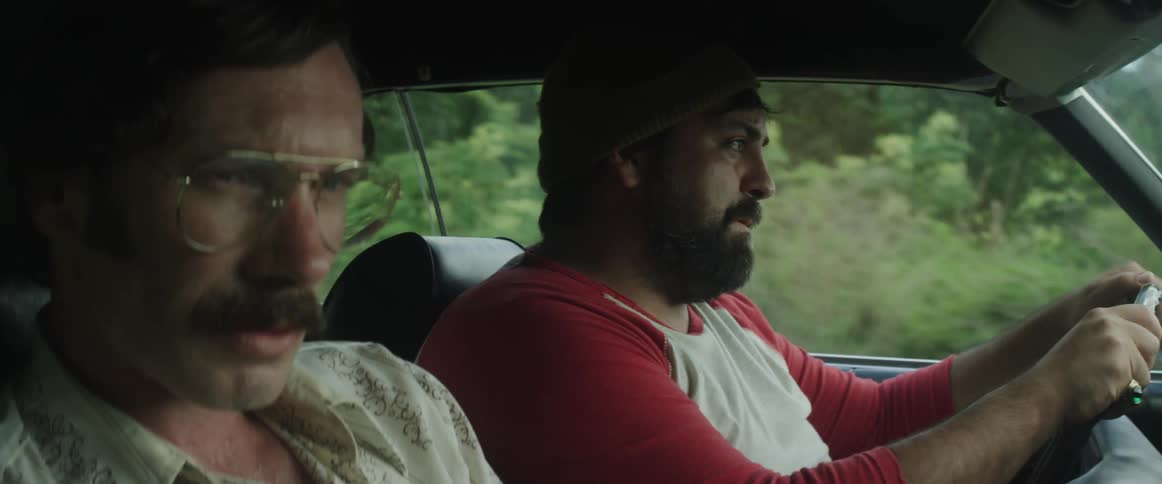 Two men sit in a car and talk as they drive past a forest area