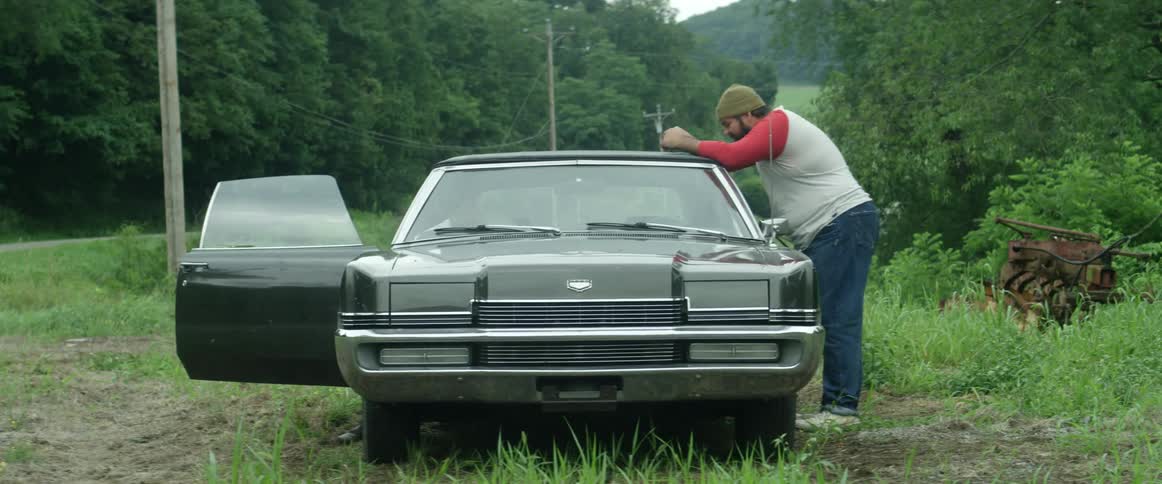 A man leans on the side of a vintage car in a rural area while another gets in the passenger side