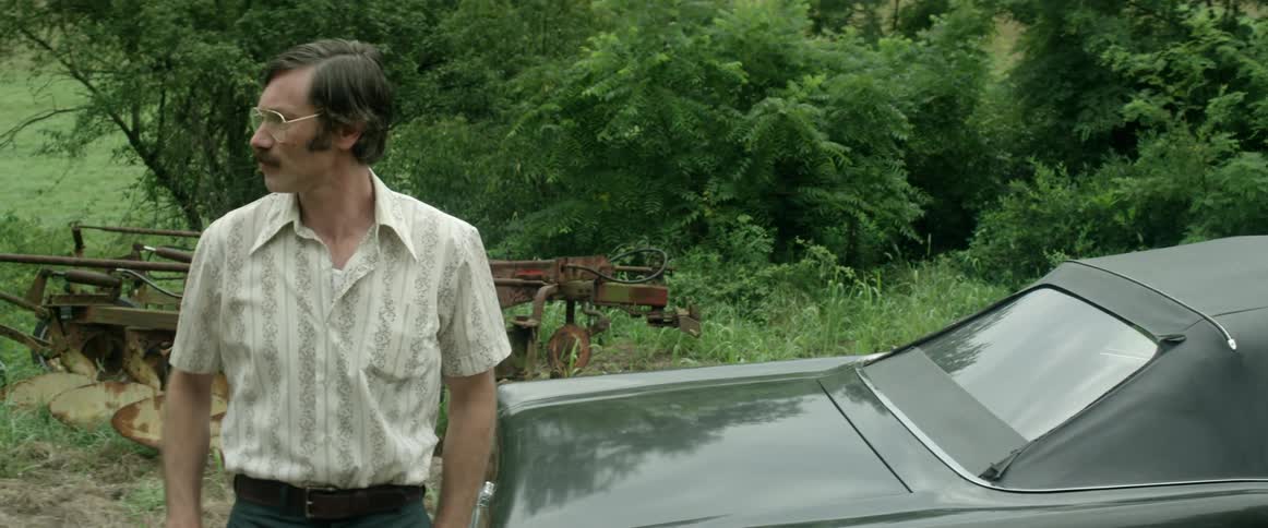A man retrieves a pistol from the trunk of his vehicle and looks around as he tucks it into the back of his pants before leisurely walking towards the front of the vehicle
