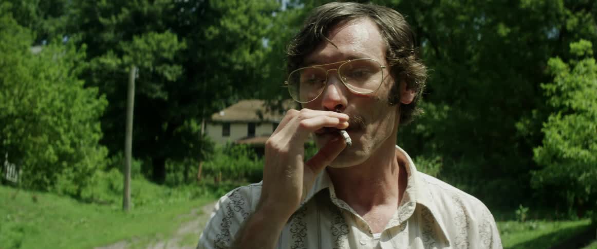 Man wearing eyeglasses talks and smokes with large house nested among trees