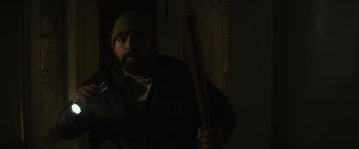 A man carrying a baseball bat shines a flashlight as he walks through a dark house