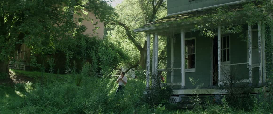 Two people standing by a house surrounded by plants with trees in the background